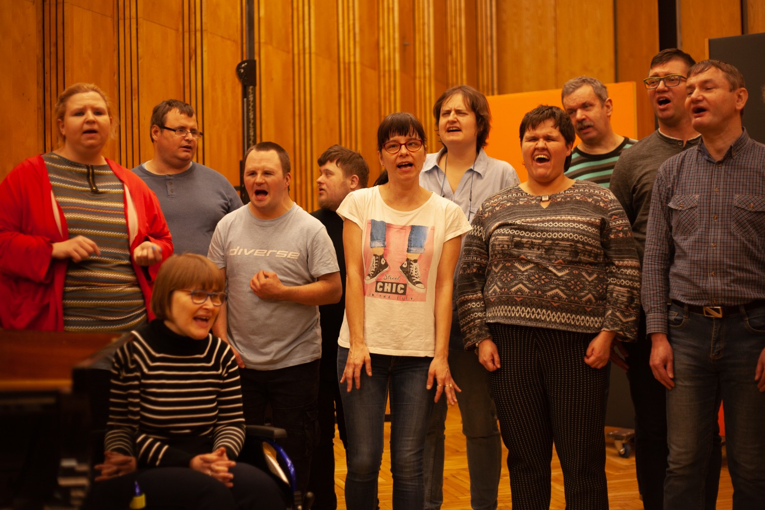 Dziesięć osób z chóru Współgłosy stoi w rzędzie i śpiewa, jedna osoba śpiewa, siedząc w wózku inwalidzkim. Próba odbywa się w radiowym studio