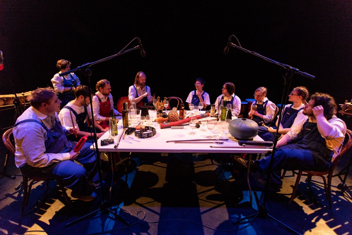 Zespół Remont Pomp gra koncert. Dziesięć osób w białych koszulach i niebieskich fatuchach siedzi przy wielkim stole, na którym są instrumenty
